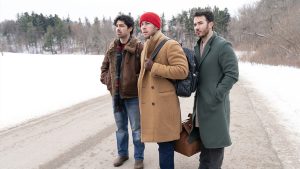 In an image from Disney+’s A Very Jonas Christmas Movie, from left to right, Joe, Nick, and Kevin Jonas stand on a snow-covered road, surrounded by trees. All are wearing heavy coats; Nick is wearing a beanie, and both Nick and Kevin are carrying bags. The trio is looking at something off in the distance, to the left.