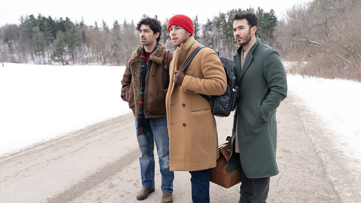 In an image from Disney+’s A Very Jonas Christmas Movie, from left to right, Joe, Nick, and Kevin Jonas stand on a snow-covered road, surrounded by trees. All are wearing heavy coats; Nick is wearing a beanie, and both Nick and Kevin are carrying bags. The trio is looking at something off in the distance, to the left.