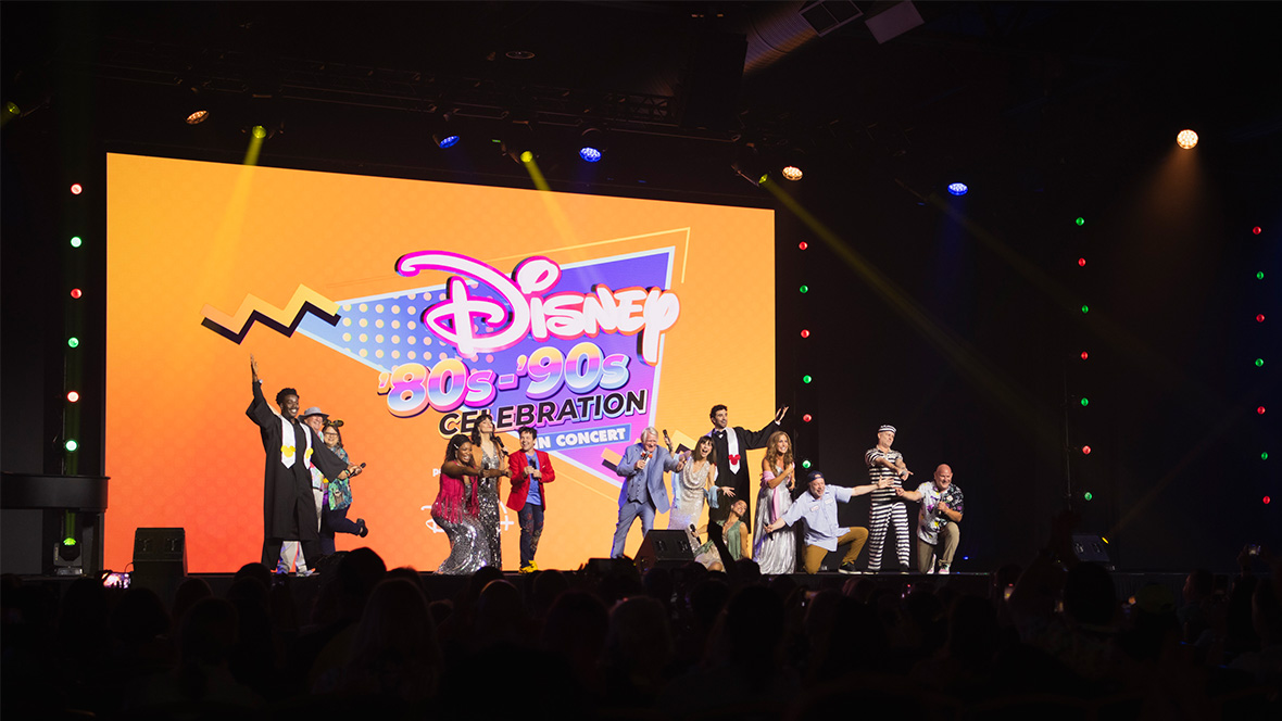 More than a dozen performers, in groups of three or more, perform onstage at Destination D23. Many have their arms out or raised as if this is the finale of a song. Behind them is an orange and purple graphic that reads: “Disney’80s-’90sCelebration in Concert.” 