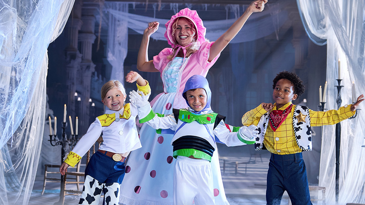A woman dressed in a Little Bo Peep (Toy Story) costume is dancing around with three children dressed in a Jessie, Buzz Lightyear, and Woody costume. They are in a spookily decorated ballroom draped with tattered white curtains and eerily lit candles.