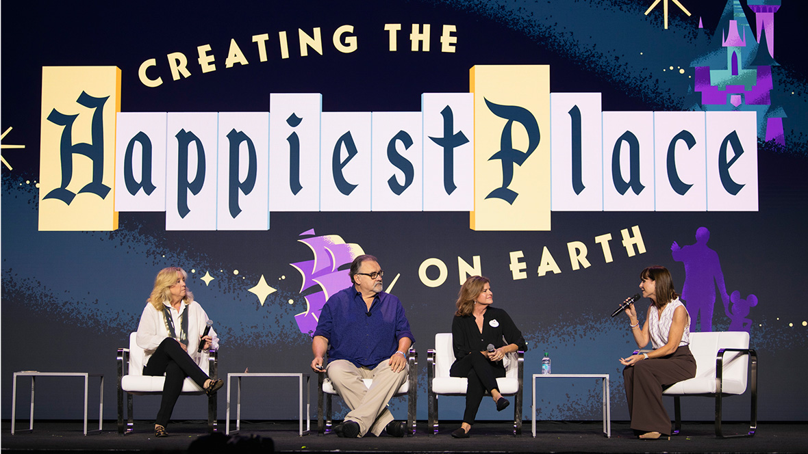 Onstage at Destination 23 are four people, seated in a row of white chairs with low tables between them. The panelists include Leslie Iwerks, on the left, and Disney Legend Don Hahn, second from left, and two additional women on the right. Behind them in Disneyland-style typography is a projection of a graphic that reads “Creating the Happiest Place on Earth.”