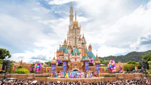 An image of Hong Kong Disneyland’s Sleeping Beauty Castle during the day, decorated for a 20th anniversary celebration. A large crowd stands at its base, and a show is happening in front of it.