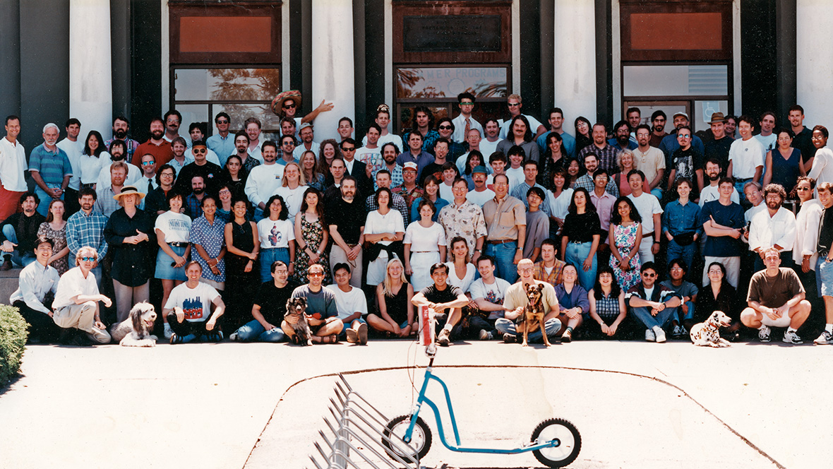 The Toy Story crew at the Point Richmond studio. Several people sitting on the ground in the front row have dogs at their sides. There is a blue scooter parked in the foreground of the photo.