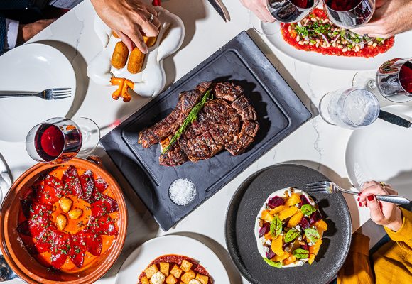 We are looking at a top-down view of a white linen-covered table with many food dishes on various plates, with hands reaching in. Dishes include a serving of steak, a pear salad, and more. Two of the hands in the table are “cheersing” with their wine glasses, filled with red wine.
