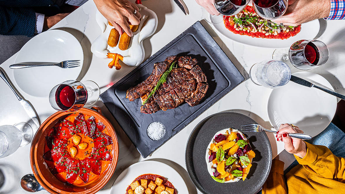 We are looking at a top-down view of a white linen-covered table with many food dishes on various plates, with hands reaching in. Dishes include a serving of steak, a pear salad, and more. Two of the hands in the table are “cheersing” with their wine glasses, filled with red wine.