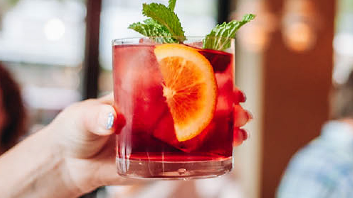 A close-up shot of a person’s hand holding a refreshing cocktail glass full of ice and dark red sangria. An orange slice floats amid the ice, while a bit of green garnish sticks out of the glass.