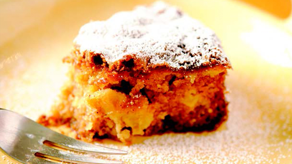 A close-up image of a plain white ceramic plate displays a squarish slice of moist apple cake, the brown crumb holding together chunks of apple. The top of the cake is dusted with powdered sugar, which is also seen on the surrounding plate. On the left, a fork lies on the plate next to the cake.