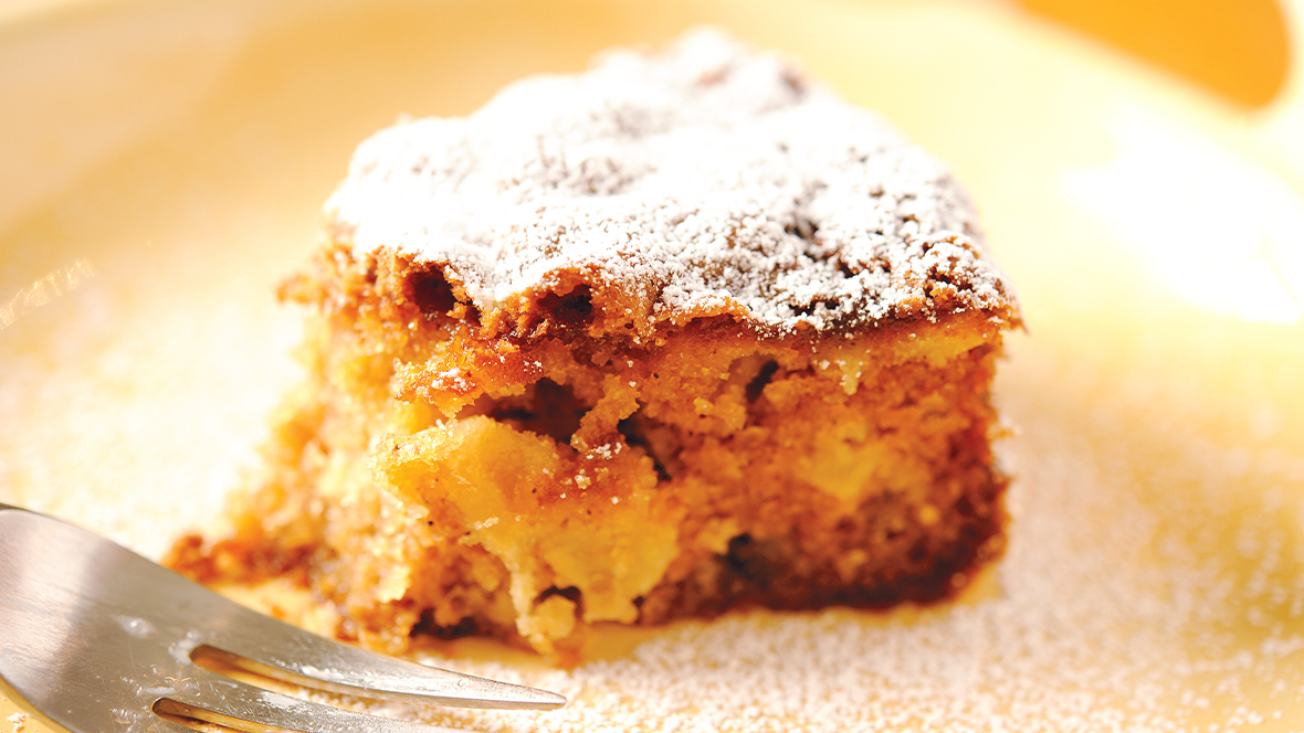 A close-up image of a plain white ceramic plate displays a squarish slice of moist apple cake, the brown crumb holding together chunks of apple. The top of the cake is dusted with powdered sugar, which is also seen on the surrounding plate. On the left, a fork lies on the plate next to the cake.