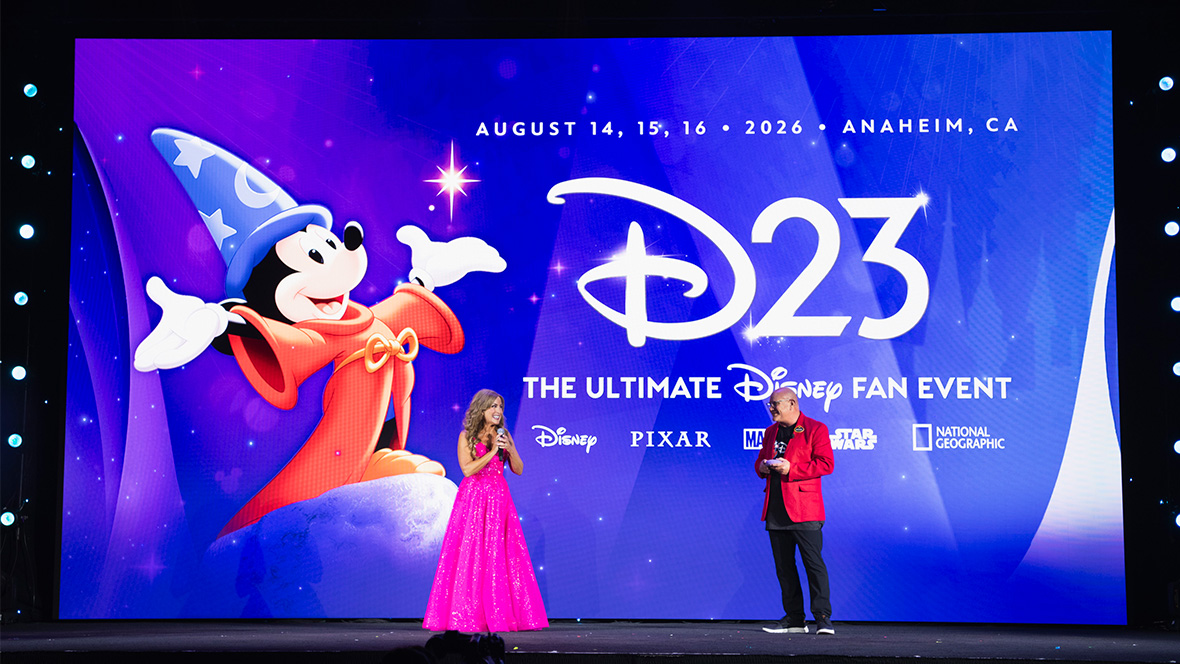 Disney Legend Jodi Benson and Michael Vargo, the head of D23, stand onstage at the Destination D23 event. Behind them is a huge projection of the graphic for D23: The Ultimate Disney Fan Event, with Sorcerer Mickey on the left and text on the right that reads: August 14, 15, and 16, 2026, Anaheim, California, D23: The Ultimate Disney Fan Event, followed by a lineup of the logos for Disney, Pixar, Marvel, Star Wars, and National Geographic. Jodi is wearing a floor-length pink gown. Michael wears a red sport jacket over a black shirt with dark pants.