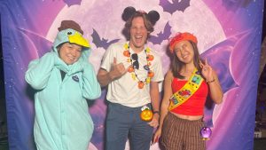 Disney cast members in costume pose in front of a Vampirina: Teenage Vampire photo-op at D23’s Halloween Boo-nanza event in Glendale, CA. The cast member on the left is wearing a Perry the Platypus-inspired onesie; the one in the middle is wearing a classic Mickey Mouse Ear hat with a jack-o-lantern and candy garland; and the one on the right is wearing a Fireside Girls-inspired scout costume from Phineas and Ferb. The backdrop features large purple bat wings against a glowing moon.