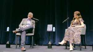 (Right): Author Rick Riordan, joined by Lisa Cabello, onstage at D23 and Disney Publishing’s “An Evening with Rick Riordan” event in New York City. The speakers sit in raised gray chairs, and in between them, a table displays a book from the Percy Jackson and the Olympians series.