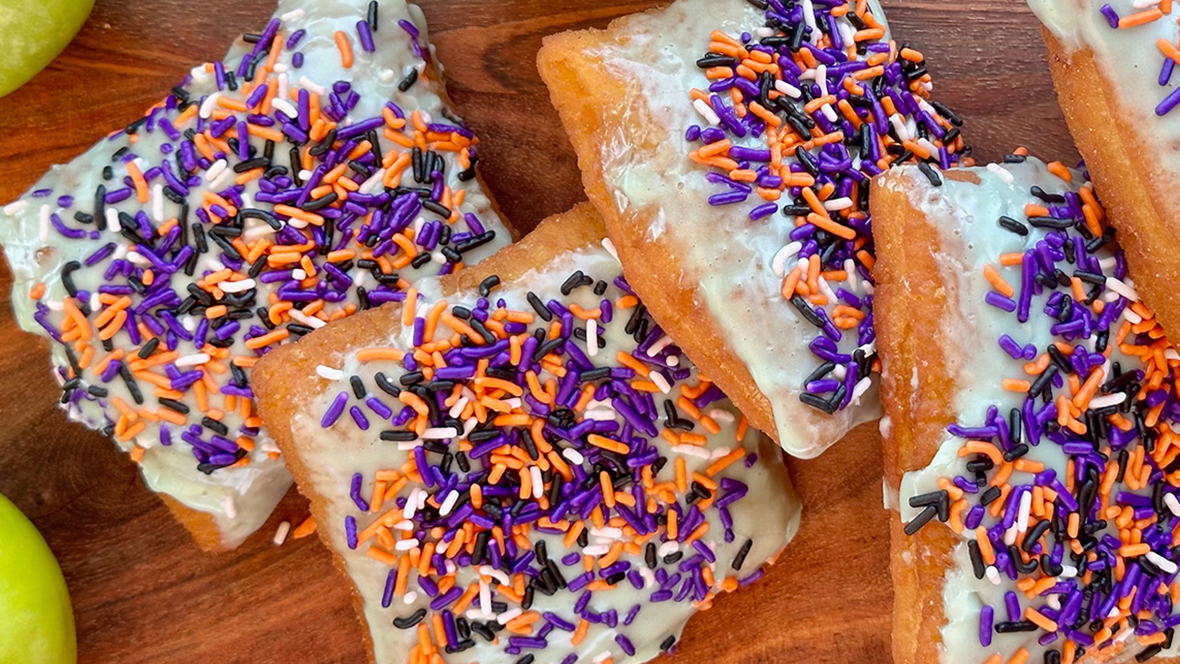 Four Caramel Apple Beignets from Beignets Expressed are topped with candied apple icing, caramel drizzle, and spooky sprinkles and served on a wooden tabletop.