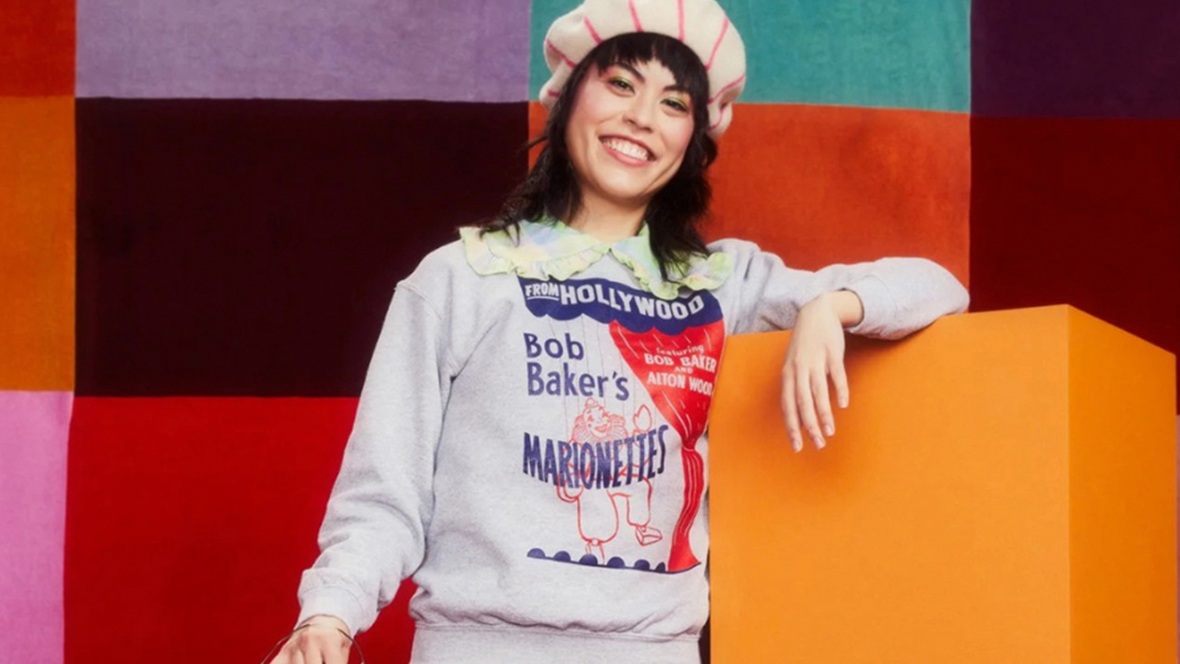 A woman is standing in front of a multicolored background wearing a Bob Baker’s Marionettes pullover and a cream and pink striped hat.