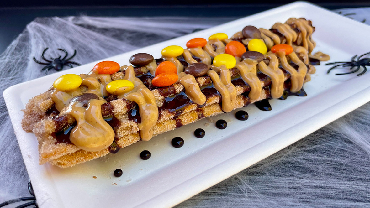 The Spooky Churro from California Churro is topped with peanut butter and chocolate sauce and topped with peanut butter candy pieces. The churro is served on a white plate and set on top of a black table covered in white faux cobwebs and faux spiders.