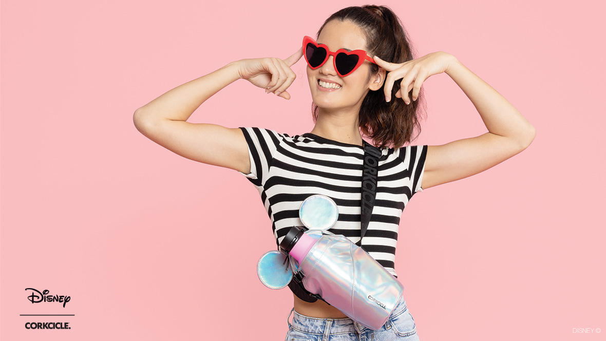 A woman in a black-and-white striped shirt points to the pair of red, heart-shaped sunglasses she is wearing. Draped over her left shoulder is a prismatic-colored Corkcicle crossbody water bottle sling bag. The top of the sling bag features a pair of Mickey Mouse ears. The woman stands in front of a light pink background. In the bottom left corner of the graphic are the Disney and Corkcicle logos, separated by a thin black line.