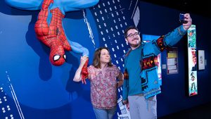 Two fans take a selfie with a statue of Spider-Man hanging upside down. The statue is being showcased by a spotlight and our web-slinging hero is holding up the &ldquo;peace&rdquo; sign. The backdrop of the photo up is blue-and-white paint drawings, simulating New York skyscrapers.