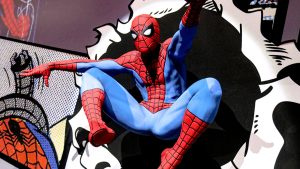 A life-size flying swinging Spider-Man statue bursts through a wall, leaping into action. The wall behind him is covered in comic book panels. The edges of the hole that our hero has swung through replicate torn paper, suggesting the hero has burst through a comic book and into the real world.