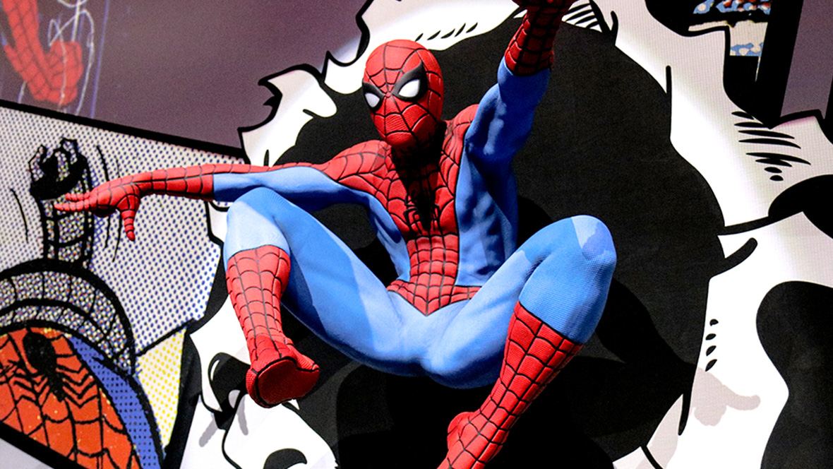 A life-size flying swinging Spider-Man statue bursts through a wall, leaping into action. The wall behind him is covered in comic book panels. The edges of the hole that our hero has swung through replicate torn paper, suggesting the hero has burst through a comic book and into the real world.