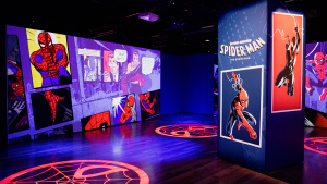 A view of the Spider-Man exhibition interior, where multiple images from Spider-Man comic books are being showcased. This includes both the Peter Parker and the Miles Morales Spider-Man throughout different eras. The ground is spotlighted with big circular red Spider-Man mask logos.