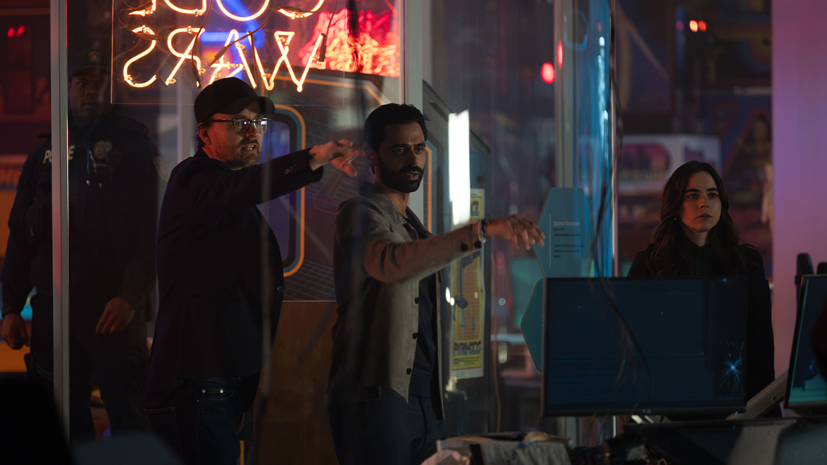 In a behind-the-scenes image from Disney’s Tron: Ares, director Joachim Rønning (left) is standing on set with actor Hasan Minhaj (middle), who portrays Ajay Singh. They are standing in what looks like an old-school office, and some neon lettering is hanging above Rønning; we see it as backwards, but it reads “Code Wars.” Several other crew members and actors can also be seen, including a woman to Minaj’s right.