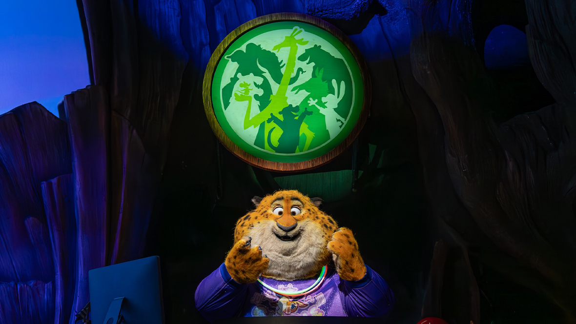 An image of the new Benjamin Clawhauser Audio-Animatronics figure for the Zootopia: Better Zoogether! show at Disney’s Animal Kingdom Theme Park. He’s sitting behind a desk and console of some kind, including a computer monitor. Above him is a Tree of Life-style logo featuring other Zootopia characters.