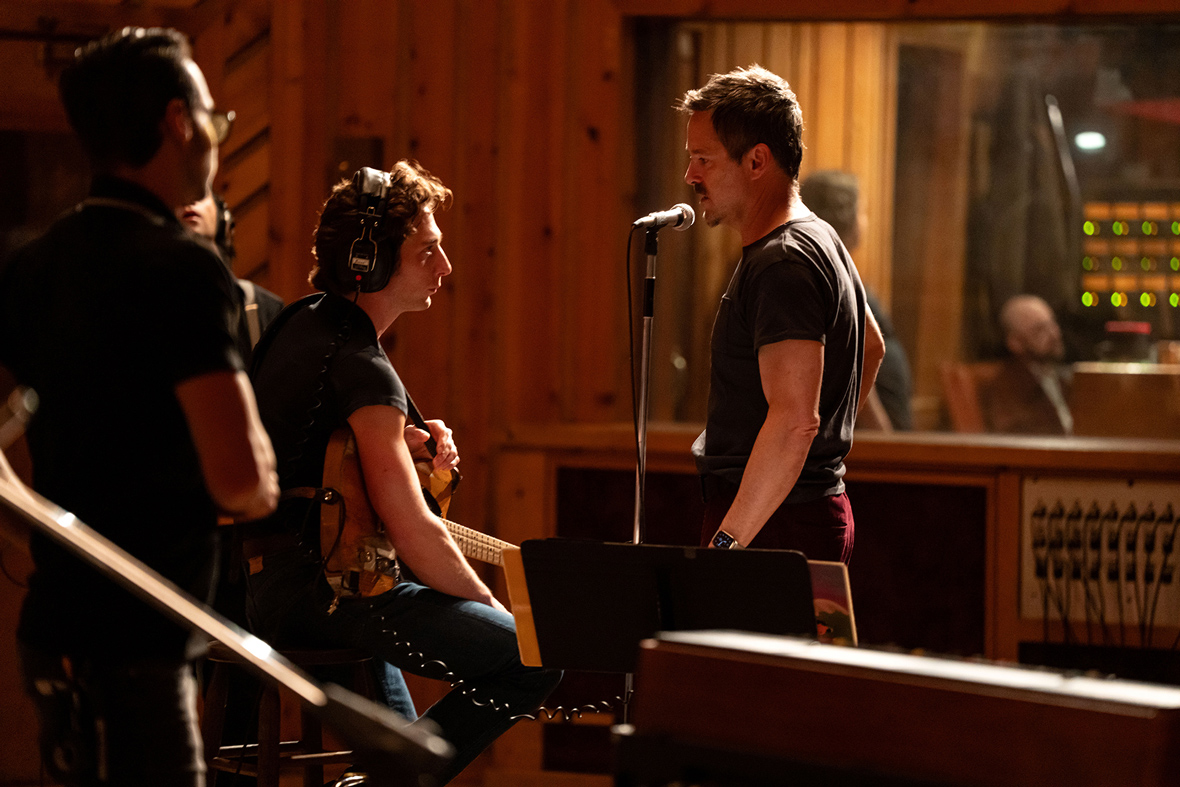Jeremy Allen White and Scott Cooper are seen in a still from 20th Century Studios’ Springsteen: Deliver Me From Nowhere. White, center left, is wearing headphones and holding a guitar; Cooper, center right, is wearing a black t-shirt and standing near a microphone on a stand.
