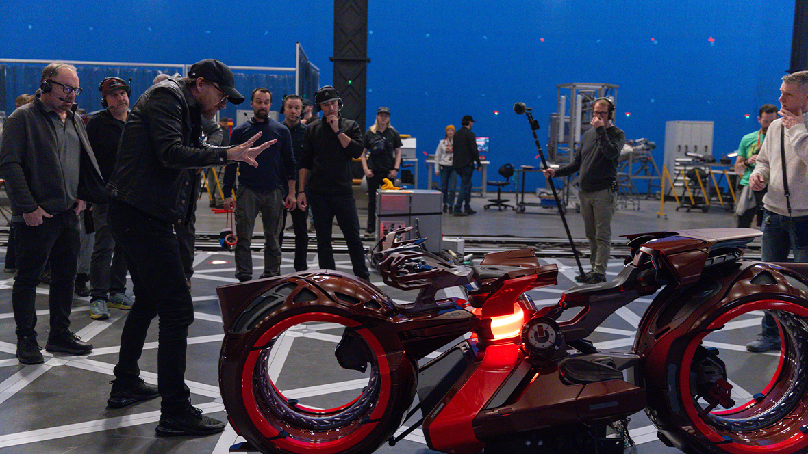 In a behind-the-scenes image from Disney’s TRON: Ares, director Joachim Rønning is seen standing to the left of a Lightcycle and surrounded by many other crew members from the film. He is gesturing toward the Lightcycle, which sits on a black-and-white grid on the floor. A large blue backdrop can be seen in the distance.