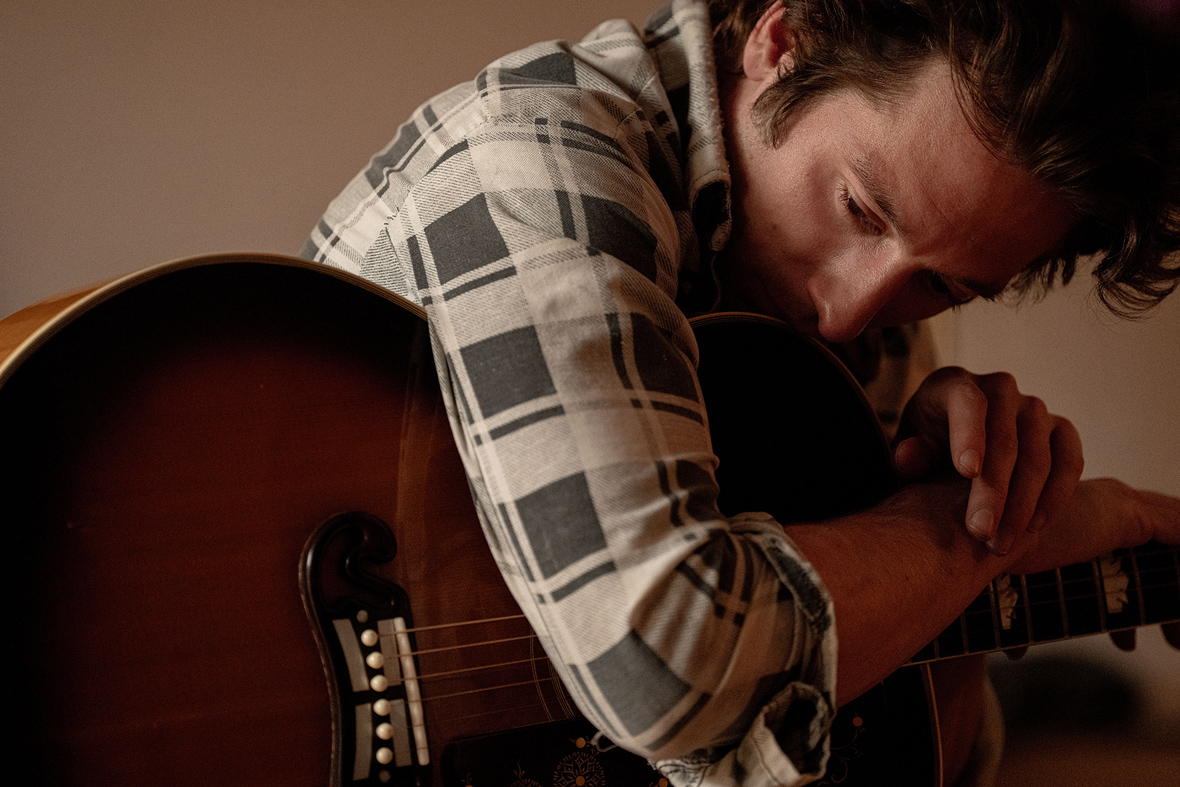 Jeremy Allen White as Bruce Springsteen in a still from 20th Century Studios’ Springsteen: Deliver Me from Nowhere. He is wearing a grey plaid shirt and holding a guitar, leaning over it with his right arm.