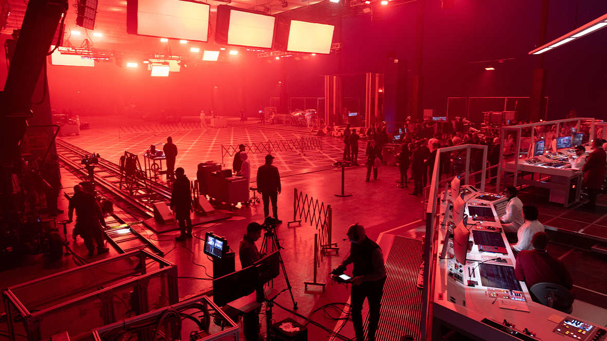 In a behind-the-scenes image from Disney’s Tron: Ares, a large soundstage is bathed in red light. Many crew members are seen working in the foreground and in the distance, and actors sitting at “workstations” are seen to the right. 