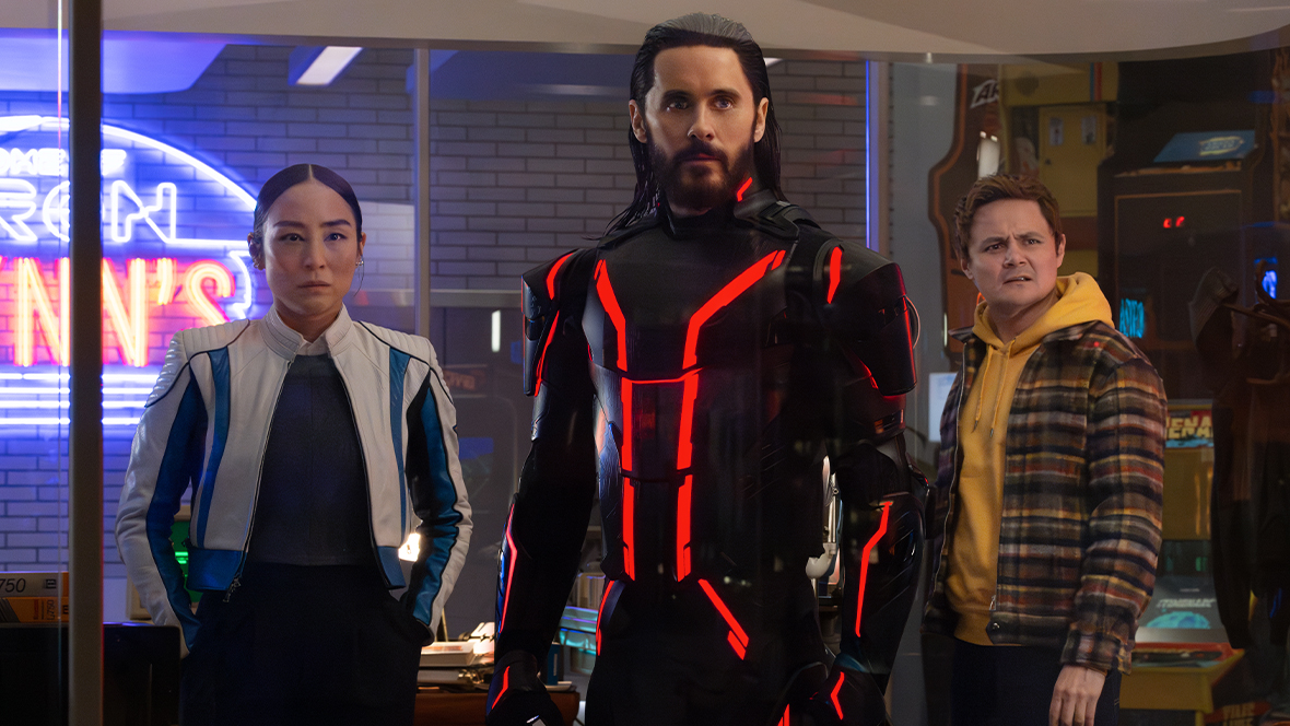 In an image from Disney’s Tron: Ares, Eve Kim (Greta Lee, left), Ares (Jared Leto, middle), and Arturo Castro (Seth Flores, right) are standing and looking at something off camera to the right. A neon sign behind Eve and somewhat outside of the image can be seen to partially say the words “Tron” and “Flynn’s,” as in Flynn’s Arcade.