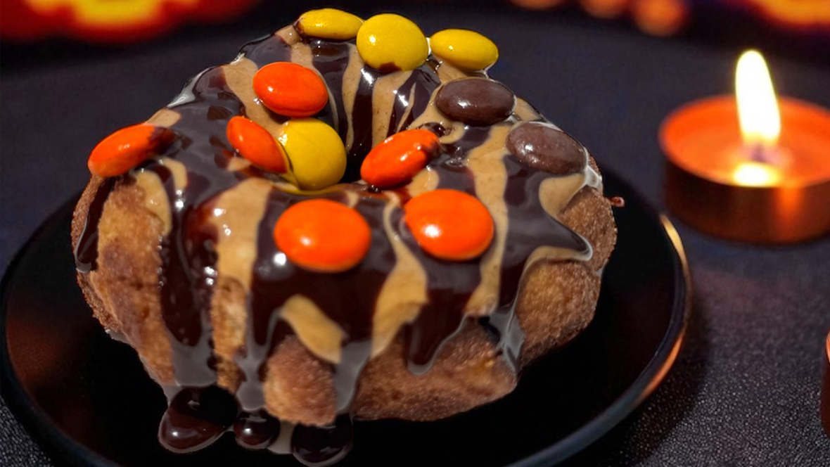 The Trick or Treat Churro Donut from Sunshine Donuts is rolled in chocolate sugar, drizzled with peanut butter and chocolate sauce, and topped with peanut butter chocolate candy pieces. The donut is served on a black plate and surrounded by lit orange candles.