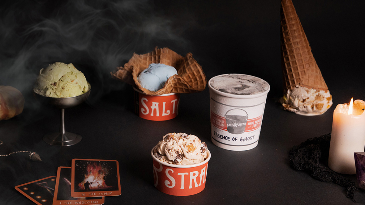 Salt & Straw’s 5 Halloween flavors, served in a variety of cups and bowls, sit on a black tabletop against a black backdrop. From left to right, they are Creepy Crawly Critters, Double Bubble Toil & Trouble, The Great Candycopia, Essence of Ghost, and Jack O’Lantern Pumpkin Bread. The table is scattered with candles, cards, and pumpkins.