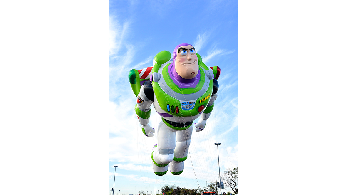 A full image of the new (2025) Buzz Lightyear balloon from the Macy’s Thanksgiving Day Parade, from the top of his head all the way down to his feet. He is posed as if flying through the air, with his wings outstretched and his hands down by his waist. A blue sky with clouds is seen behind him.