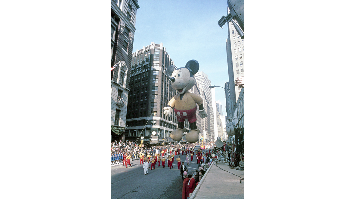 A historical image of the Mickey Mouse balloon from the 1974 Macy’s Thanksgiving Day Parade. Mickey is wearing a yellow shirt and red shorts with white buttons, and his handlers are also wearing outfits of yellow and red.