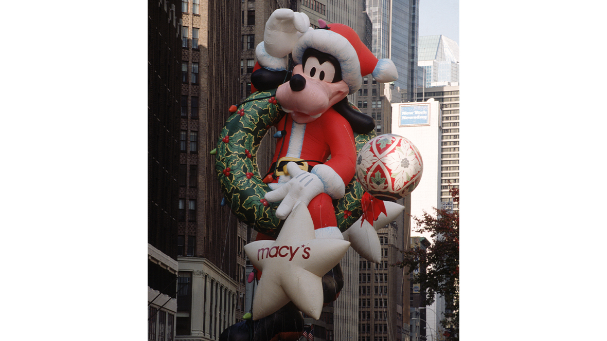 A historical image of the Santa Goofy balloon from the 1992 Macy’s Thanksgiving Day Parade. Goofy has a Santa hat and coat on and has a wreath with holiday lights slung over one arm. A smaller balloon in front of him is shaped like a star and features the Macy’s logo.