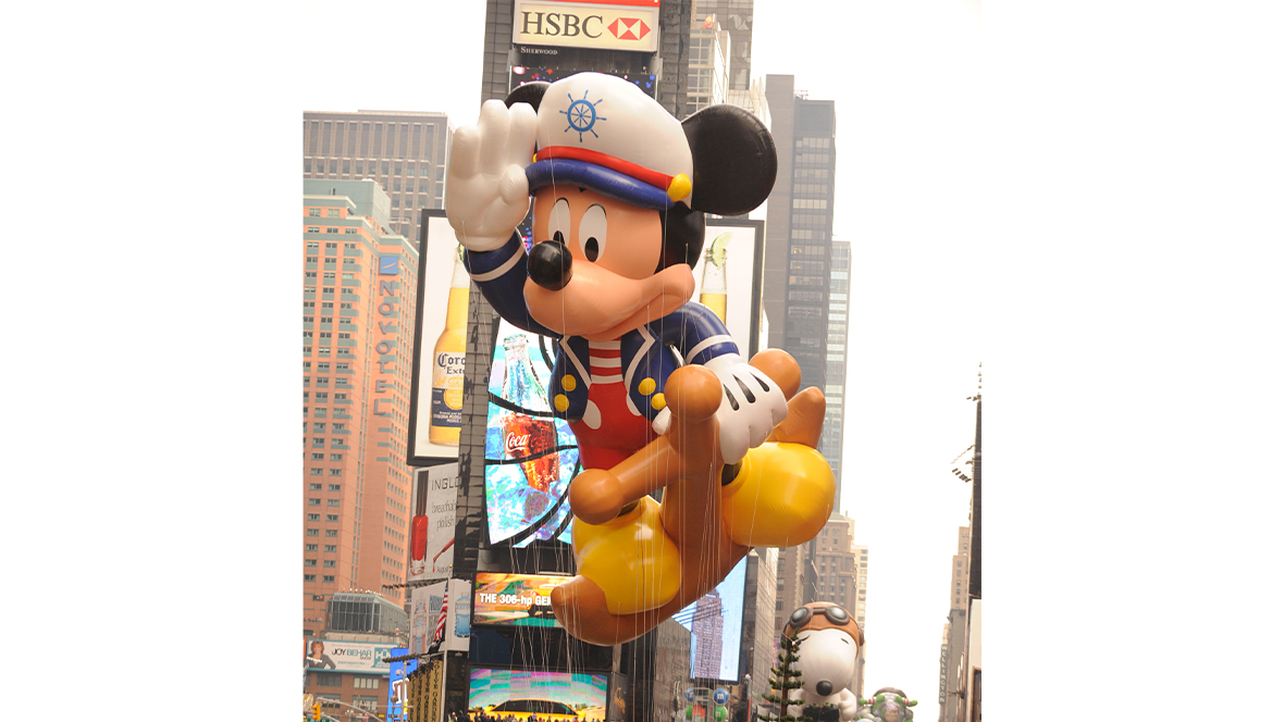 A historical image of the Sailor Mickey balloon, celebrating Disney Cruise Line’s two new cruise ships, in the 2009 Macy’s Thanksgiving Day Parade. Mickey is wearing a red, white, and blue uniform and is saluting the crowd below.