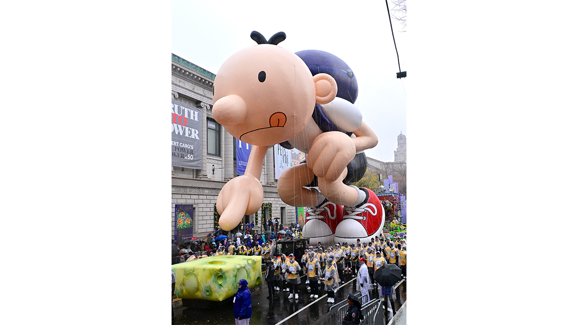 An image of the Greg Heffley (from Diary of a Wimpy Kid) balloon in the Macy’s Thanksgiving Day Parade. He is wearing a backpack, and in front of the balloon, on the street, is a large inflated piece of cheese, that balloon Greg is actively trying to avoid touching!