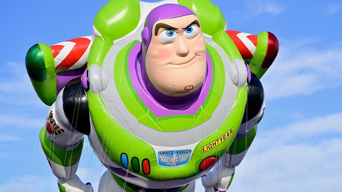 A closer image of the new (2025) Buzz Lightyear balloon from the Macy’s Thanksgiving Day Parade. He is posed as if flying through the air, with his wings outstretched and his hands down by his waist. A blue sky with clouds is seen behind him.