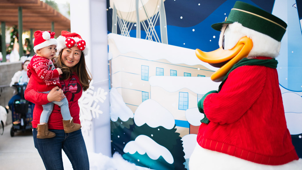 A mother holding her young child in festive red holiday outfits smiles as they meet a character dressed as Donald Duck in a green hat and red coat against a snowy, winter-themed backdrop.