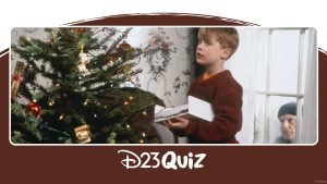 Scene from the first Home Alone film: Kevin stands beside a decorated Christmas tree holding an open box, while one of the burglars peers through the window behind him; the image is framed with a ‘D23 Quiz’ banner.