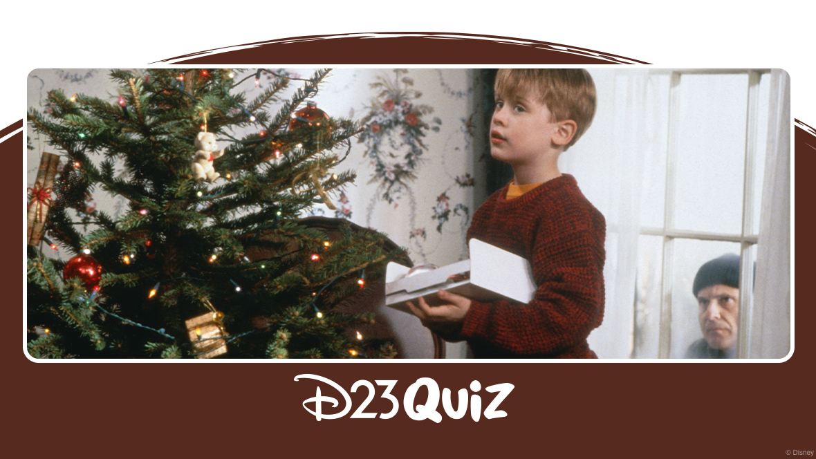 Scene from the first Home Alone film: Kevin stands beside a decorated Christmas tree holding an open box, while one of the burglars peers through the window behind him; the image is framed with a ‘D23 Quiz’ banner.