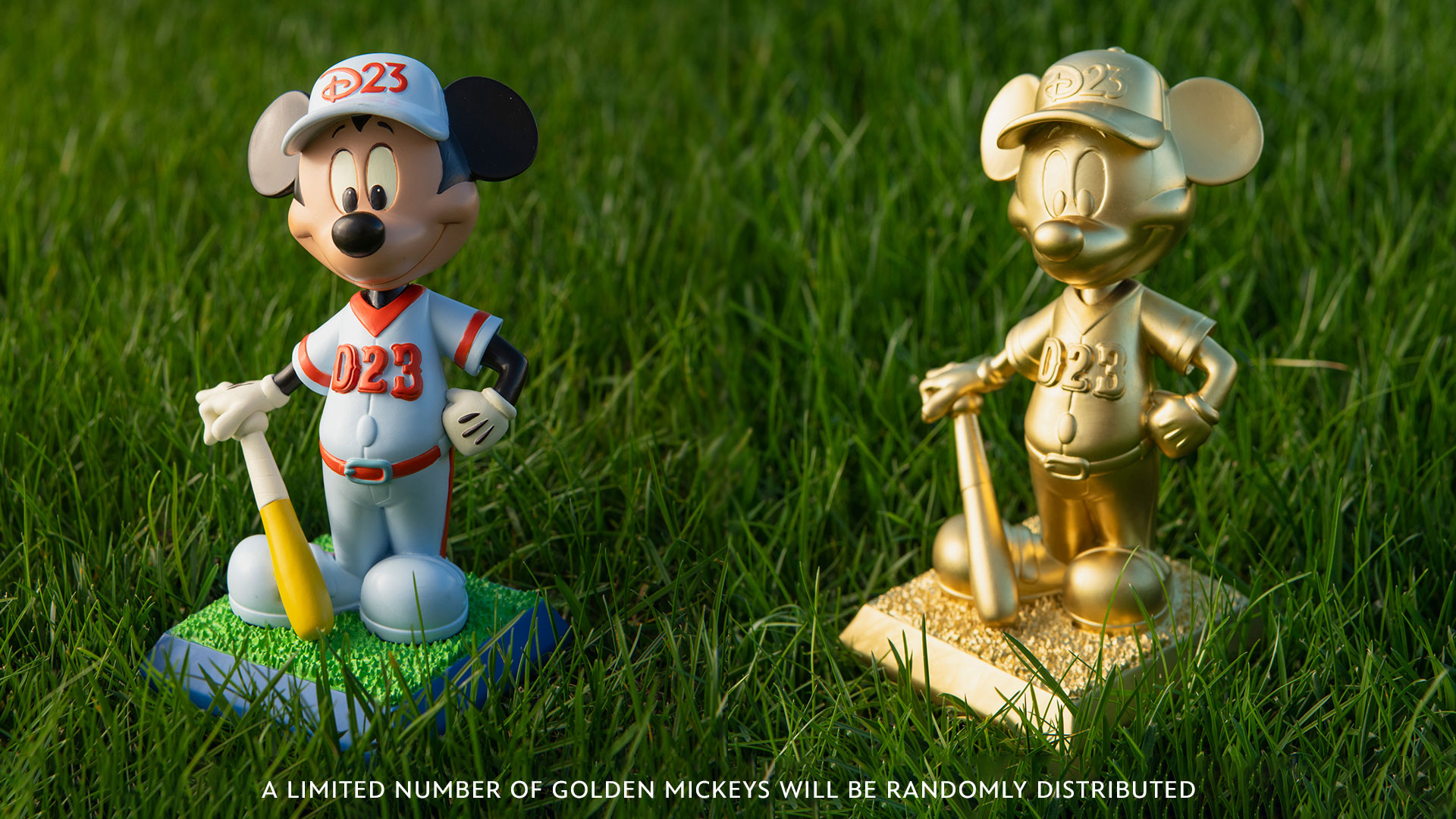 Two Mickey Mouse baseball figurines stand side by side on grass. The left figurine is in full color, wearing a blue and red D23 baseball uniform and holding a yellow bat, while the right figurine is entirely gold. Text at the bottom reads: "A limited number of golden Mickeys will be randomly distributed."