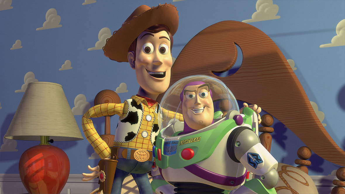 A still from 1995’s Toy Story, featuring Sheriff Woody (voiced by Tom Hanks), left, in his iconic hat and holster, and Buzz Lightyear (voiced by Disney Legend Tim Allen), with the clear hood of his spacesuit covering his head. Woody has his arm around Buzz and they are smiling at the camera, standing on the bed in Andy’s room.