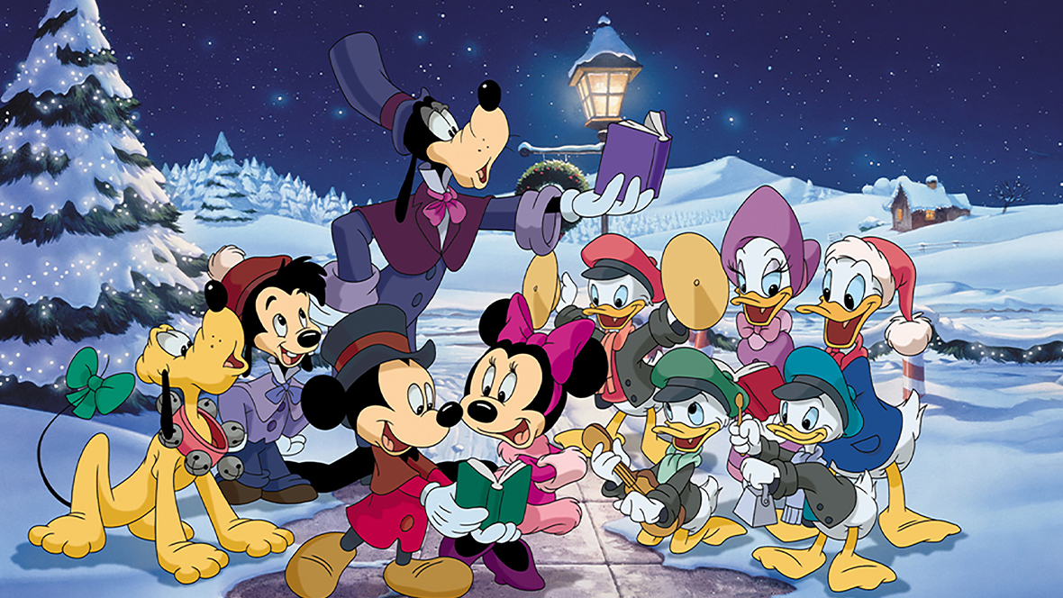A still from 1999’s animated Mickey’s Once Upon a Christmas, featuring the whole cast, including Mickey Mouse, Minnie Mouse, Donald Duck, Goofy, and Pluto. Everyone appears to be singing Christmas carols. A lit tree is to the group’s left.