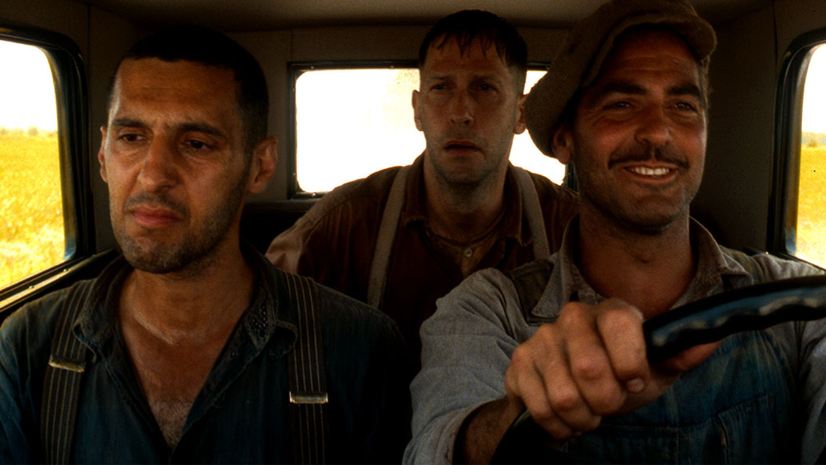 In a still from 2000’s O Brother, Where Art Thou?, Pete (John Turturro), left, Delmar (Tim Blake Nelson), middle, and Ulysses Everett McGill (George Clooney) are sitting inside a car; McGill is driving and smiling, while the two others look more serious. It’s daytime, and fields of yellow can be seen out the car’s windows.