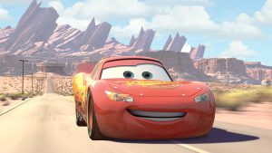 An image from the 2006 Disney and Pixar animated film Cars featuring Lightning McQueen (voiced by Owen Wilson), as he&rsquo;s driving down the highway outside of Radiator Springs, which can be seen in the distance behind him.