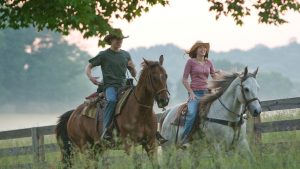 In a still from 2009&rsquo;s Hannah Montana the Movie, Travis (Lucas Till), left, and Miley (Disney Legend Miley Cyrus), right, are riding on horseback through tall grass. Both are wearing cowboy hats and are smiling.