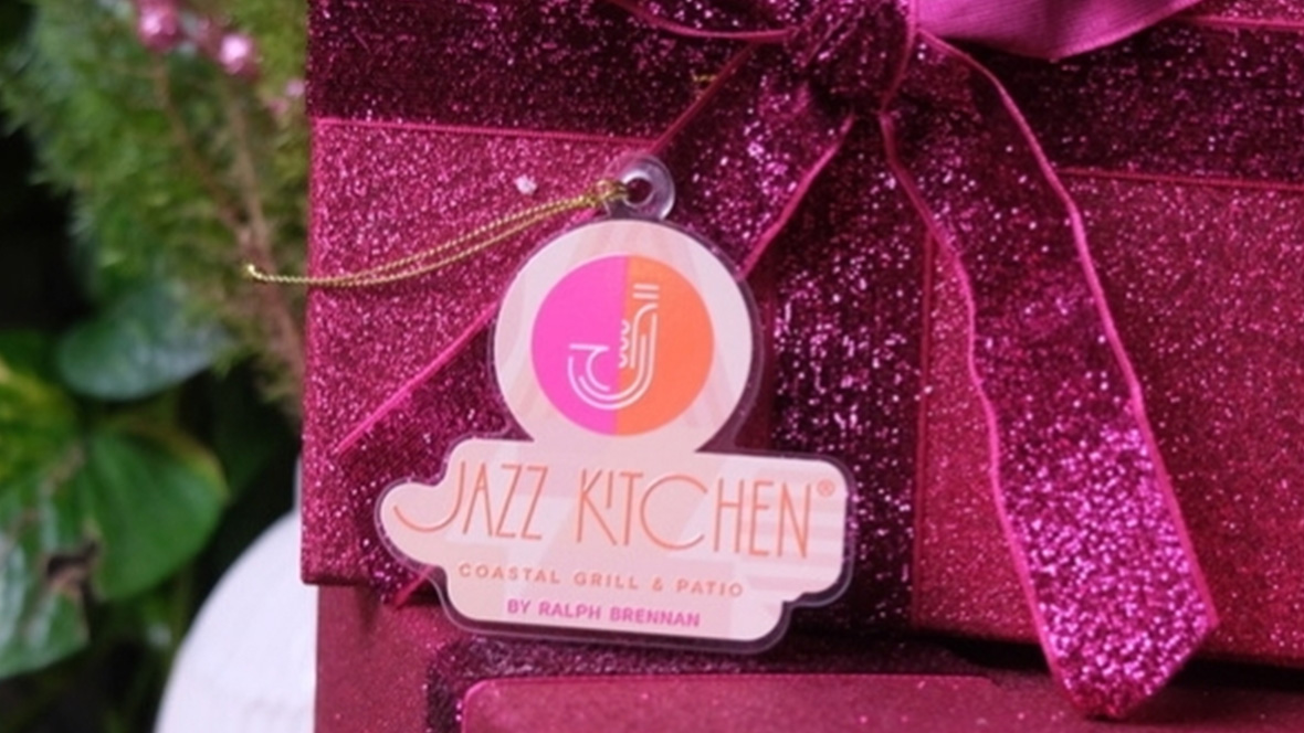 A Christmas Tree ornament sits by a gift box wrapped in sparkling pink wrapping paper, adorned with a matching bow. The ornament sports the “Jazz Kitchen Coastal Grill & Patio by Ralph Brennan” logo. The logo features a minimalist graphic of a saxophone.