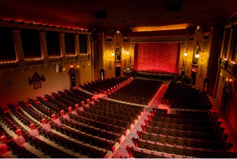 An elevated view from above of an empty auditorium in the historic Music Box Theatre. At the front of the theater, a large stage is framed by an elaborately carved proscenium and a red, scalloped curtain