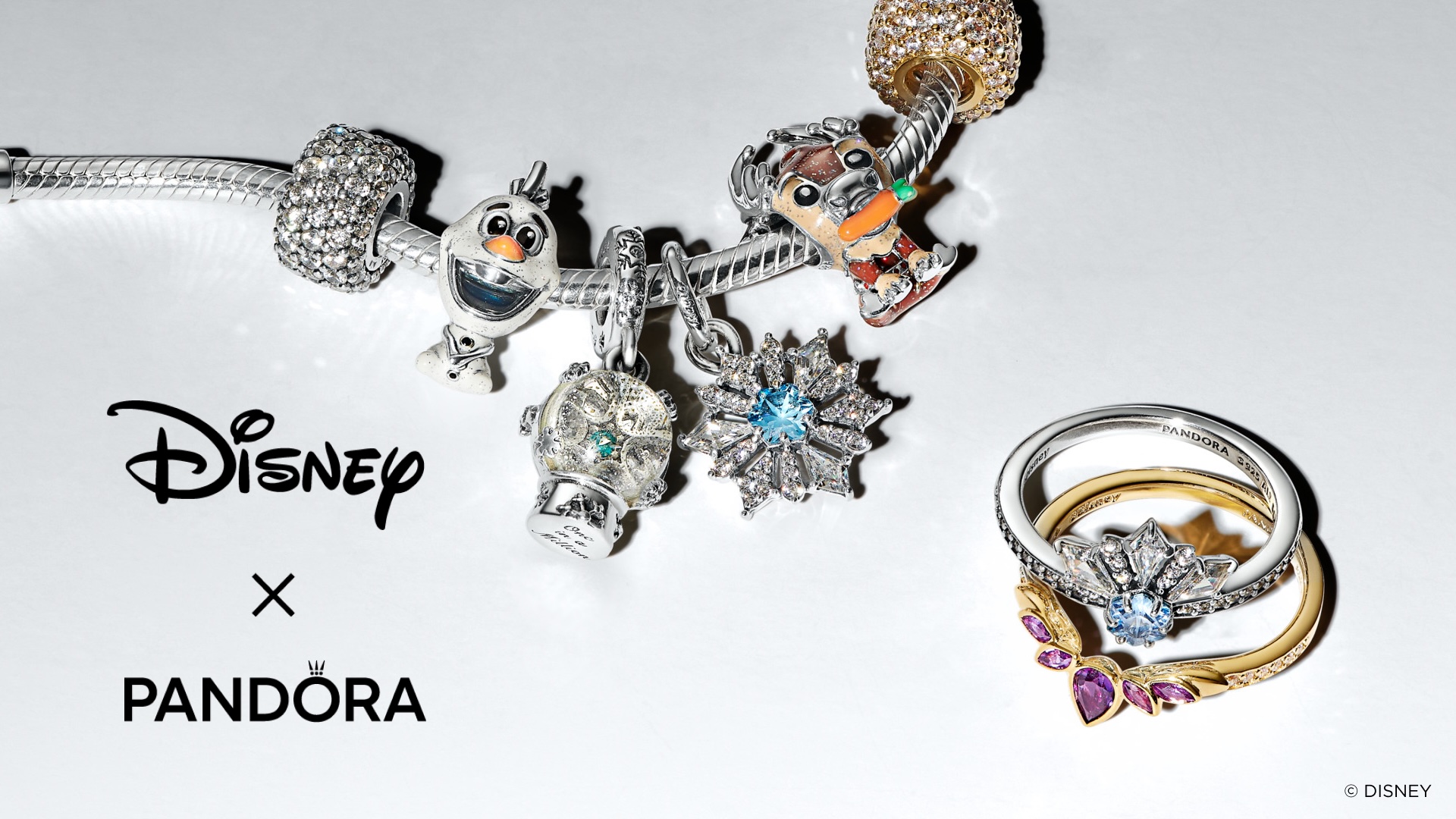 A Pandora bracelet rests against a white background with charms adorning it, themed to the Disney film Frozen and including Olaf, Sven, a snow globe, and a snowflake. There are two rings next to the bracelet, which are themed to Elsa and Anna by color. There is copy at the bottom of the graphic which reads: “Disney (logo) x Pandora (logo).”